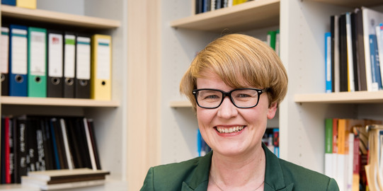 Porträt einer Frau mit Brille. Im Hintergrund Bücherregale.