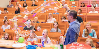 KinderUni Ein Mann schwenkt einen großen Reifen durch die Luft und produziert Seifenblasen. Mehrere Kinder sitzen im Hintergrund auf Hörsaal-Bänken und betrachten den Mann.