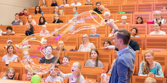 KinderUni Ein Mann schwenkt einen großen Reifen durch die Luft und produziert Seifenblasen. Mehrere Kinder sitzen im Hintergrund auf Hörsaal-Bänken und betrachten den Mann.