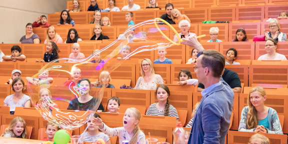 A man swings a large hoop through the air and produces soap bubbles. Several children sit on auditorium benches in the background and watch the man.