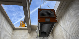 Foto: Ein großes technisches Bauteil wird mit einem Kran durch eine Öffnung in einem Betongebäude gehoben.