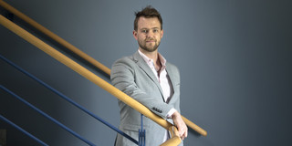 A portrait shot of a man on a staircase with a curved banister made of light wood.