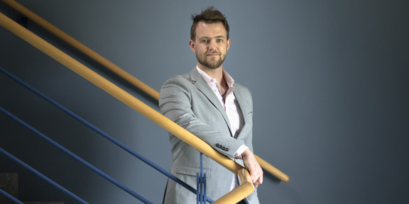 Matthias Faes Eine Portraitaufnahme eines Mannes auf einer Treppe mit einem geschwungenen Geländer aus hellem Holz.