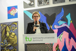 Foto: Eine Frau mit blondem kurzen Haar steht vor verschiedenen Kunstwerken an einem Pult. Sie hält ein Mikrofron in der Hand und spricht.