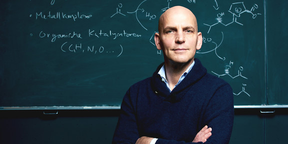 A portrait of Benjamin List in front of a blackboard with chemical formulas