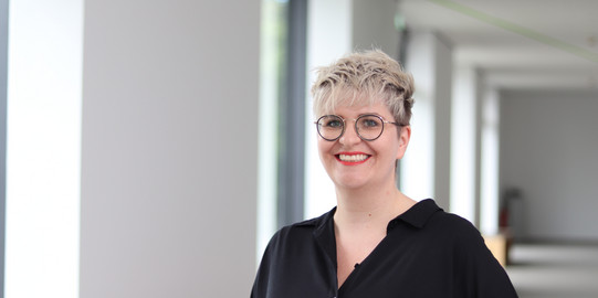 Laura Naegele Portrait einer Frau mit Brille und kurzem blonden Haar in einem hellen Flur