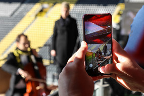 Zwei Hände halten ein Smartphone, mit dem Cellist Andrei Simion gerade fotografiert wird und der unscharf im Hintergrund zu sehen ist.