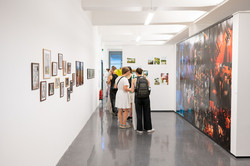 Das Bild zeigt einen breiteren Gang mit Menschen. Auf der linken weißen Wand hängen Rahmen mit Kunstwerken. Die rechte Wand ist komplett mit einem Kunstwerk versehen.