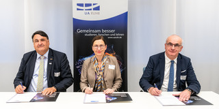 Two men and a woman are sitting at a table, each signing a document in open folders in front of you. In the background is an exhibitor with the inscription: "UA Ruhr - studying, researching and teaching better together".