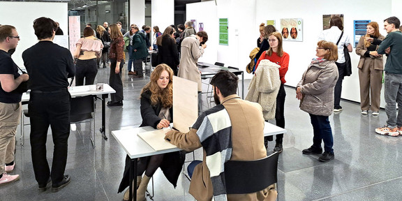 Das Bild zeigt einen Ausstellungsraum mit Besucher:innen. Im Zentrum des Bildes sitzen sich zwei Menschen an einem Tisch gegenüber. Auf dem Tisch selbst steht eine Trennwand. Die Personen haben ihre eigenen Unterarme jeweils auf dem Tisch auf der einen und der anderen Trennseite aufgelegt. Im Hintergrund sind weitere Tische und Besucher:innen zu sehen sowie eine weiße Wand mit Leinwänden.
