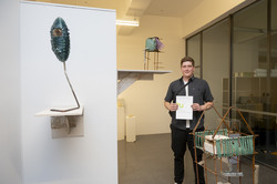 Rundgang Kunst Kunstpreis Plastik Ein Student mit Urkunde steht zwischen zwei Plastiken auf Wandregalen links und einer stehenden Plastik rechts.