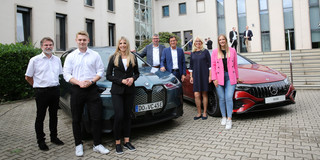 Vor einem dunkelblauen BMW-SUV und einem roten Mercedes stehen vier Männer und drei Frauen und posieren für ein Foto.