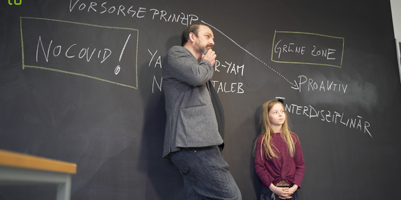 KinderUni Corona Ein Mann und ein Mädchen stehen mit dem Rücken vor einer Tafel