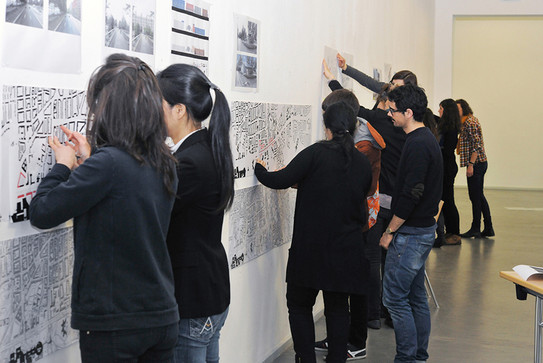 Eine Gruppe von Studierenden schaut sich Baupläne an, die an einer Wand hängen.