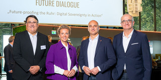 A woman and three men are standing in front of a presentation displayed in large format in the background.
