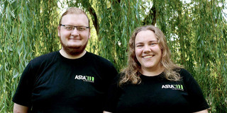 AStA head officers 2024 Two people wearing black shirts with the AStA logo and standing in front of a green willow: Darius Weitekamp on the left, Lara Witte on the right
