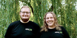 AStA-Vorstand 2024 Zwei Personen tragen schwarze Shirts mit AStA-Logo und stehen vor einer grünen Weide: Links Darius Weitekamp, rechts Lara Witte