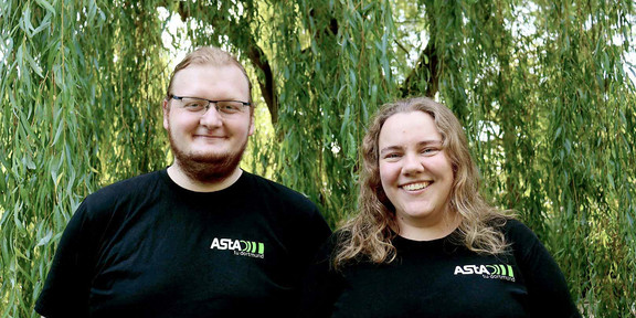 AStA-Vorstand 2024 Zwei Personen tragen schwarze Shirts mit AStA-Logo und stehen vor einer grünen Weide: Links Darius Weitekamp, rechts Lara Witte