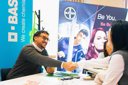 An einem Messestand schütteln ein Firmenvertreter (links) und eine internationale Studentin (rechts) über dem weißen Tisch Hände und lächeln. Auf dem Tisch liegen Firmenmaterialien, im Hintergrund sind verschiedene Firmenbanner zu sehen.