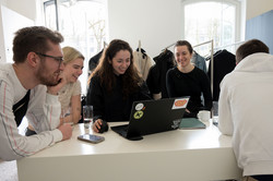 Eine Gruppe von vier Studierenden lachen und schauen an einem Gaderobentisch an einem Laptop. Rechts steht ein Student mit dem Rücken zur Kamera vor dem Gaderobentisch.
