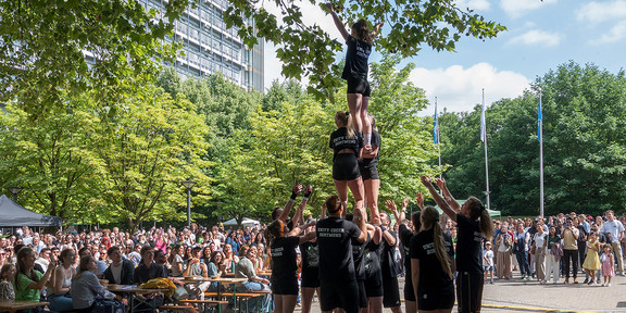 Das Cheerleading-Team der TU Dortmund zeigt eine Pyramidenfigur auf dem Martin-Schmeißer-Platz. Im Hintergrund ein großes Publikum an Picknick-Tischen und im Stehen.