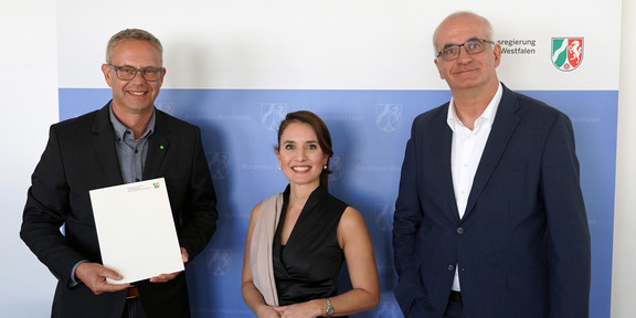 Foto: Eine Frau in schwarzem Kleid und zwei Männern in Anzüge mit Brillen stehen vor einem Banner der Landesregierung Nordrhein-Westfalen. Der linke Mann hält eine Urkunde in der Hand.