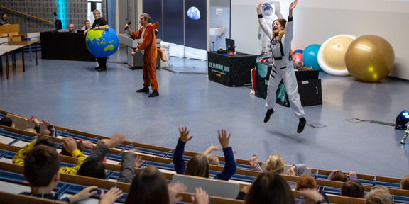 Drei Personen stehen während der DLR_Raumfahrt_Show im vollbesetzten Audimax. Sie tragen Kostüme einer Erde, eines Kängurus und einer Astronautin. Die Astronautin springt in die Luft.