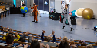 Three people stand in the packed Audimax during the DLR_Space_Show. They wear costumes of an earth, a kangaroo and an astronaut. The astronaut jumps into the air.