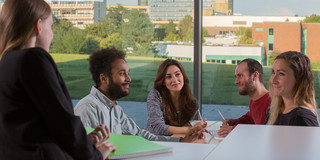 Five students sit at a table with pens in their hands and paper in front of them on the table.