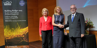 Drei Personen stehen auf einer Bühne bei der Tagung der Academia Europaea in Barcelona. Prof. Susanne Fengler steht in der Mitte und wird von Präsident Prof. Donald Dingwell auf der rechten Seite und Vize-Präsidentin Prof. Eva Kondorosi auf der linken Seite begrüßt.