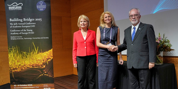 Drei Personen stehen auf einer Bühne bei der Tagung der Academia Europaea in Barcelona. Prof. Susanne Fengler steht in der Mitte und wird von Präsident Prof. Donald Dingwell auf der rechten Seite und Vize-Präsidentin Prof. Eva Kondorosi auf der linken Seite begrüßt.