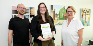 Foto: Drei Personen stehen gemeinsam in einem Büro; die mittlere Person hält ein Zertifikat in den Händen.