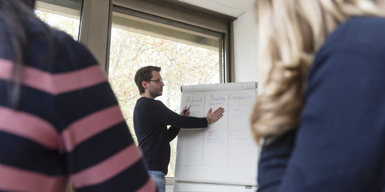 Dozent ist zwischen zwei Studentinnen zu sehen und schreibt etwas auf ein Board