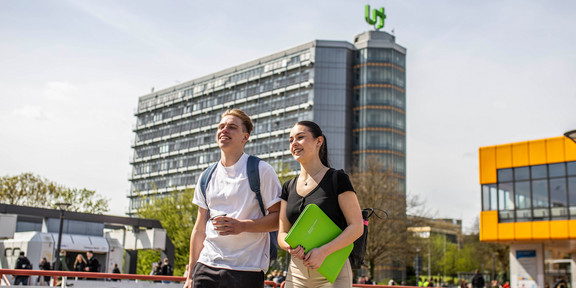 Zwei Studierende laufen über den Campus der TU Dortmund. Hinter ihnen ist der Mathe-Tower und die Mensa.