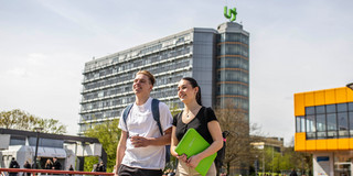 Zwei Studierende laufen über den Campus der TU Dortmund. Hinter ihnen ist der Mathe-Tower und die Mensa.