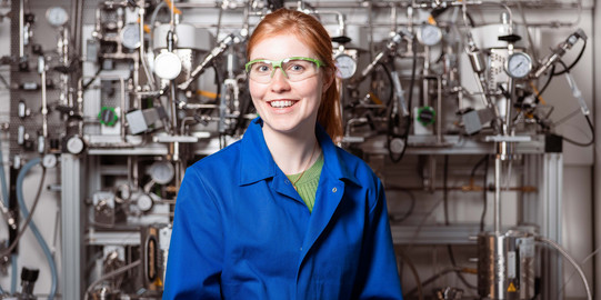 Junge Frau in blauem Arbeitsanzug steht vor technischen Apparaturen.
