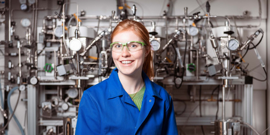 Young woman in blue work suit stands in front of technical equipment.