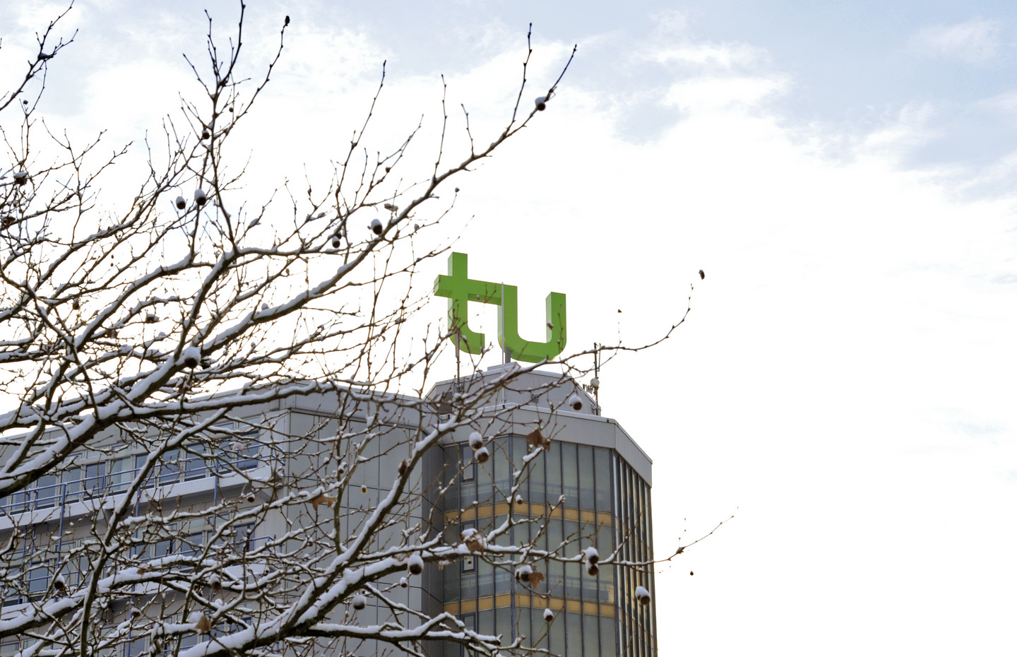Die obere Hälfte des Mathetowers mit grünem TU-Logo an einem sonnigen Wintertag. Auf der linken Seite ragen schneebedeckte Äste eines Baumes ins Bild.