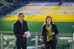 Auf dem Foto sieht man links einen Mann stehen, der ein Mikrofon trägt und Moderationskarten in der Hand hält. Rechts steht eine Frau, die in ein Mikrofon spricht und einen Schal mit dem TU Dortmund und BVB Logo trägt.