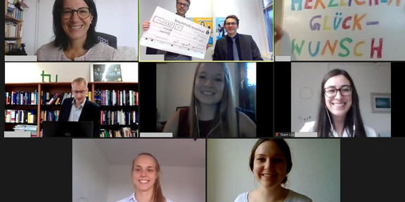 Video conference with eight participants. One participant holds a "Congratulations" sign into the camera, another the check with the prize money.