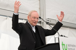 A man at the lectern raises both arms upward.