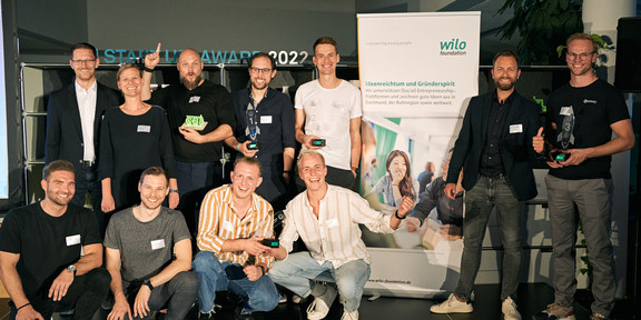 The TU Start-up Award jury Dr. Martin Oettmeier, Prof. Tessa Flatten and Nic Lecloux stand with the start-up teams next to a banner from the wilo Foundation.