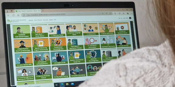 Das Bild zeigt einen Laptopbildschirm mit mehreren Kacheln, der unten rechts zum Teil von der Schulter einer davorsitzenden Person verdeckt wird.