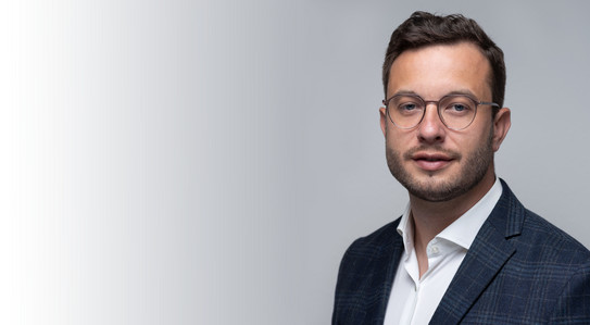 Porträtfoto von Prof. Sascha von Behren vor hellgrauem Hintergrund: Prof. Behren trägt einen Anzug und Brille.