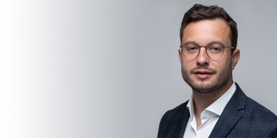 Porträtfoto von Prof. Sascha von Behren vor hellgrauem Hintergrund: Prof. Behren trägt einen Anzug und Brille.