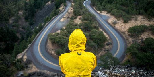 Wechseln_Straße Person in einer gelben Jacke sitzt auf einen Felsen und blickt auf eine Straßen Kurve.