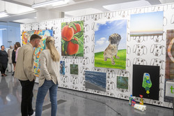 Rundgang Kunst Kunstwand An einer Wand hängen verschiedene Kunstwerke. Links im Bild sind Menschen, die sich unterhalten.
