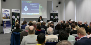Zu sehen ist ein Veranstaltungssaal mit Publikum und Prof. Martina Havenith, die vor einer Leinwand steht und eine Rede hält.