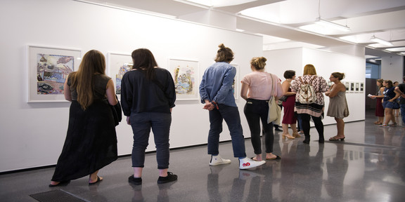 Visitors to an exhibition look at pictures hanging on a wall.