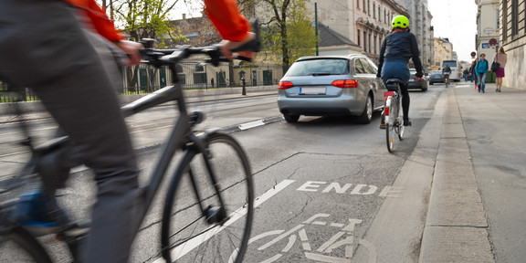 Zwei Radfahrer*innen fahren auf einer städtischen Straße. Der markierte Radweg endet. Autos fahren links neben ihnen.